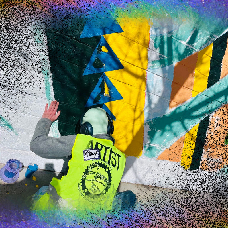 This image shows an artist in a yellow safety vest, with their back to the camera. They are squatting down in front of a textured outdoor wall, painting lifework on a mural. The mural is half finished and is made of geometric shapes in white, turquoise, blue, yellow, dark green, and coral